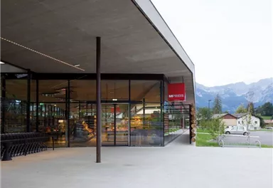 A modern shop with large glass windows and a clear, minimalist design. In the background, mountains and a rural setting are visible.