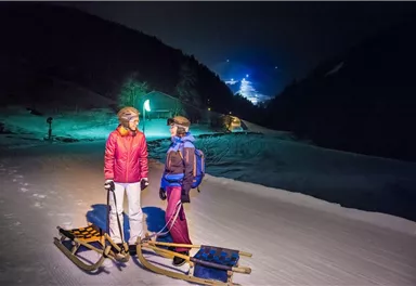 Zwei Personen stehen nachts auf einer verschneiten Piste mit Schlitten. Im Hintergrund leuchten Lichter und eine Berglandschaft ist sichtbar.