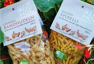 Two packages of pasta, one with ribbon noodles made from ancient spelt and the other with wheat spirals, are lying among colorful flowers. The packaging is eco-friendly and attractively designed.