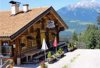 Eine traditionelle Berghütte mit Holzbauweise und einem einladenden Außenbereich. Im Hintergrund sind majestätische Berge und ein klarer blauer Himmel zu sehen.