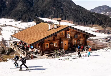 Ein gemütliches Holzhaus in den Bergen, umgeben von schneebedeckten Hängen. Skifahrer genießen die Piste vor der Hütte.