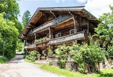 Ein traditionelles Holzhaus mit mehreren Balkonen, umgeben von grünen Bäumen und Pflanzen. Der Weg führt zum Haus und schafft eine einladende Atmosphäre.