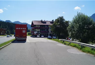 A multi-story building stands on a street, surrounded by trees and mountains. In the foreground, a red truck is parked.