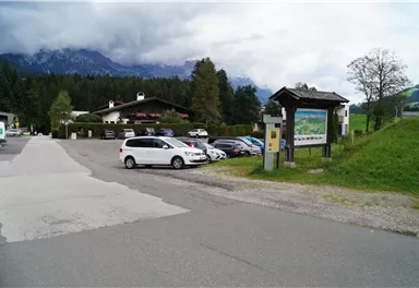 A parking lot in a picturesque setting with mountains in the background. On the left side, there is an information sign visible.
