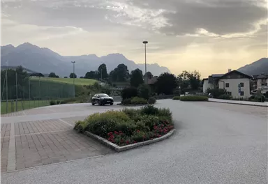 Eine ruhige Landschaft mit Bergen im Hintergrund und einer Straße. Zu sehen sind auch einige Blumenbeete und ein parkendes Auto.