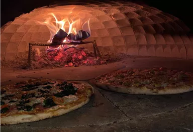 Ein traditioneller Holzofen mit loderndem Feuer und zwei Pizzen im Vordergrund. Die Pizzen sind goldbraun und knusprig.
