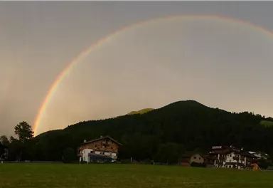 A rainbow arches over the landscape with gentle hills. In the foreground, some houses and green meadows can be seen.