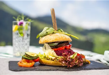 Ein appetitlicher Burger mit frischem Gemüse und einem Holzstab. Im Hintergrund ist eine grüne Landschaft und ein Getränk mit Zitronen und Blumen zu sehen.