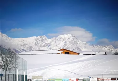 Eine schneebedeckte Landschaft mit majestätischen Bergen im Hintergrund. Ein Holzgebäude steht im Vordergrund, unter einem klaren blauen Himmel.