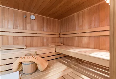 Eine gemütliche Sauna mit Holzbank und Handtuch. An der Wand hängt eine Uhr, und ein Holzfass steht im Raum.