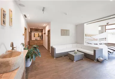Ein modernes Wohnzimmer mit einer hellen Couch und einem Holzfußboden. Im Hintergrund sind Pflanzen und Kunstwerke an der Wand zu sehen.