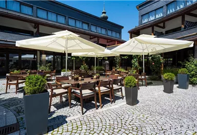 Ein einladender Außenbereich mit Holztischen und -stühlen, umgeben von Pflanzen. Große Sonnenschirme spenden Schatten an einem klaren, sonnigen Tag.