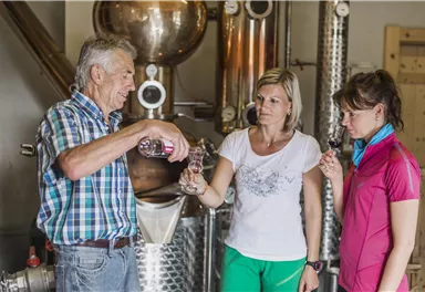 Ein älterer Mann schenkt zwei Frauen Getränke in einer Spirituosenbrennerei aus. Die Frauen beobachten interessiert und halten Gläser in der Hand.