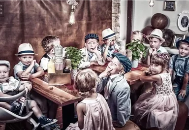 A group of children is sitting at a table in a cozy room. They are wearing elegant clothing and various hats while looking at the camera.