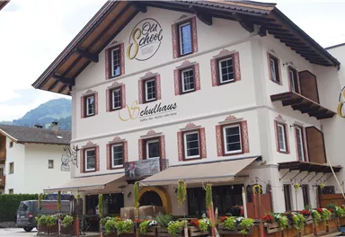 A traditional building with a balcony roof and several windows. In front of the house, there are flower boxes and a terrace with seating.