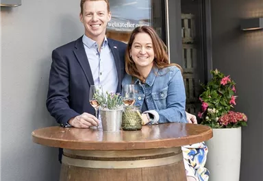 A couple stands at a wine barrel table and smiles relaxedly. In the background, blooming plants and an entryway can be seen.