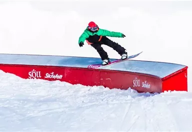 A snowboarder is performing a trick on a metal ramp in the snow. In the background, snowy trees and a bright sky can be seen.