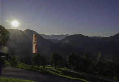 A picturesque mountain landscape with the rising sun in the background. In the foreground, a flag is waving between the trees.