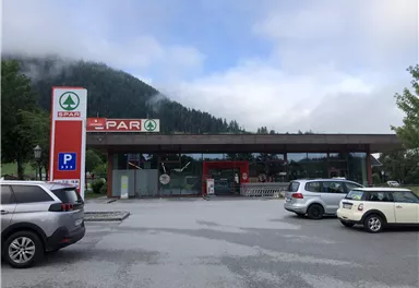 Ein SPAR-Laden in einer ländlichen Umgebung, umgeben von Bergen und Bäumen. Vor dem Geschäft steht ein Parkplatz mit mehreren Autos.