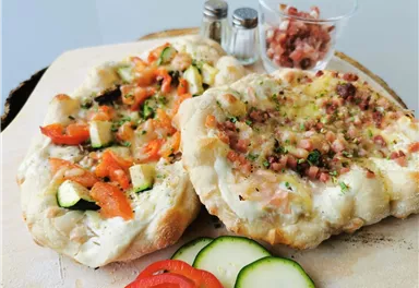 Two hearty Flammkuchen on a wooden board, one with vegetables and the other with ham. Fresh vegetables and spices complete the dish.