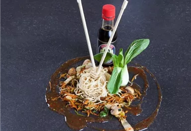 Eine Schüssel mit asiatischen Nudeln, umgeben von frischem Gemüse. Ein Fläschchen Sojasauce und Essstäbchen ergänzen das Gericht.