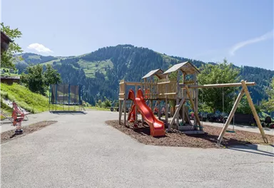 Ein Spielplatz mit einer Rutsche und einem Trampolin im Freien. Im Hintergrund sind grüne Berge und ein klarer blauer Himmel zu sehen.