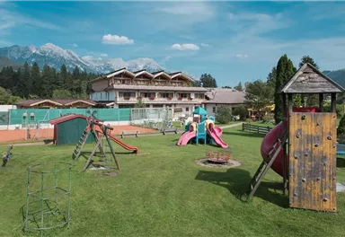 A playground with various equipment and a grassy area. In the background, mountains and a building are visible.