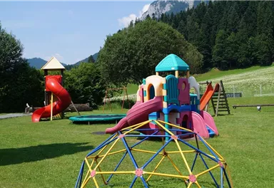 A playground with colorful play equipment and slides. In the background, green meadows and mountains can be seen.