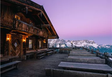 A rustic wooden house with a terrace that offers a view of the mountains. The sky is colored in soft shades of purple and pink.