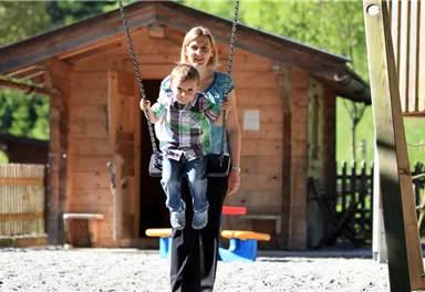 Eine Frau steht auf einem Spielplatz hinter einem schwingenden Kind. Im Hintergrund ist ein Holzhaus und eine grüne Umgebung zu sehen.