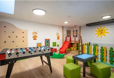 Ein bunter Spielraum für Kinder mit einem Kicker, einer Rutsche und einer Kletterwand. Die Wände sind mit fröhlichen Motiven dekoriert, und es gibt farbige Sitzgelegenheiten.