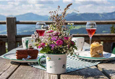An idyllic table with a flower arrangement, cake, and glasses of rosé wine. The surroundings showcase a picturesque mountain landscape.