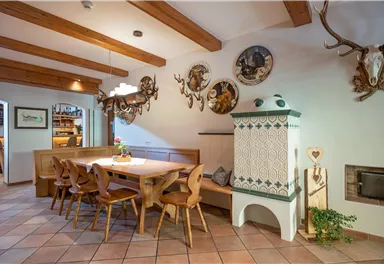 Ein einladender Essbereich mit einem langen Holztisch und Stühlen. An den Wänden hängen dekorative Jagdtrophäen und eine traditionelle Ofenheizung ist sichtbar.