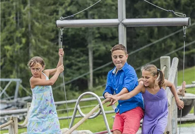 Drei Kinder spielen auf einer Holzplattform im Wasser. Sie lachen und haben Spaß in einer schönen, grünen Umgebung.