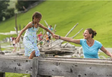 Eine Mutter hält die Hand ihrer Tochter, während sie über einen Holzsteg balanciert. Im Hintergrund sind grüne Wiesen und spielende Kinder zu sehen.