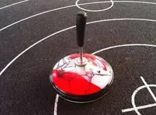 Ein kunterbunter Kreisel steht auf einem Sportplatz mit schwarzen Linien. Der Boden hat verschiedene Spielfeldmarkierungen.