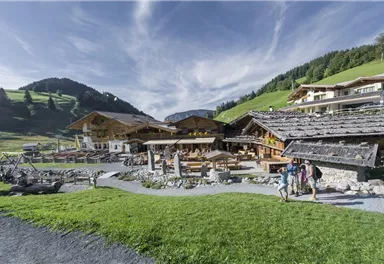 A picturesque alpine inn surrounded by green meadows and gentle hills. Three hikers are walking on a path that leads to the entrance.