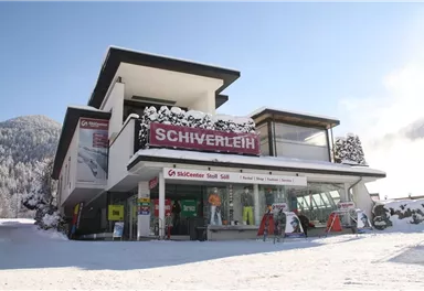 Ein modernes Geschäft für Skiverleih im verschneiten Winterlandschaft. Die Fassade zeigt große Fenster und ein einladendes Schild mit der Aufschrift "Schiverleih".