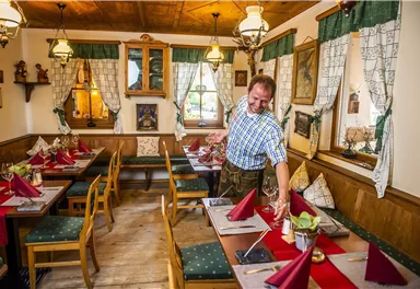 Ein gemütliches Restaurant mit traditionellen Holzmöbeln und einer einladenden Atmosphäre. Ein Bediener deckt Tische mit roten Servietten und Gläsern.