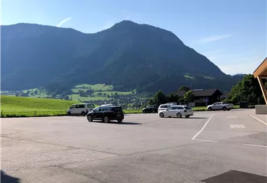 A parking lot with several cars and a view of an impressive mountain landscape. The sky is clear and blue, and the green of the meadows is clearly visible.