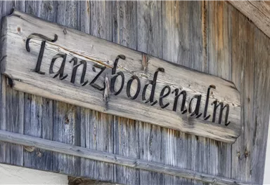 A wooden sign with the inscription "Tanzbodenalm". It is located on a rustic wall.