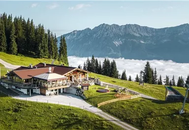 A picturesque mountain cabin surrounded by green meadows and tall trees. In the background, majestic mountains rise above a sea of fog.