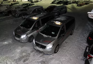 Zwei silberne Taxis stehen auf einem verschneiten Parkplatz. Die Fahrzeuge sind teilweise mit Schnee bedeckt und es sind weitere Autos im Hintergrund sichtbar.