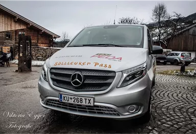 Ein silbernes Taxi mit dem Schriftzug „Sölleuken Pass“ steht auf einer gepflasterten Straße. Im Hintergrund ist ein Holzgebäude und eine ländliche Umgebung zu sehen.