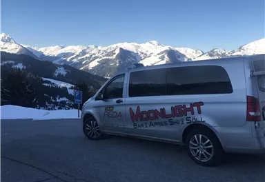 Ein silberner Van steht auf einem Parkplatz mit schneebedeckten Bergen im Hintergrund. Die Umgebung ist winterlich und sonnig.