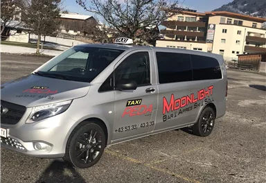 Ein silbernes Taxi mit der Aufschrift "Moonlight" steht auf einem Parkplatz. Im Hintergrund sind Bäume und Gebäude zu sehen.