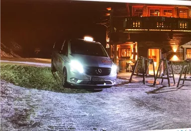 Ein Mercedes-Fahrzeug steht auf einem schneebedeckten Weg bei Nacht. Im Hintergrund ist ein beleuchtetes Gebäude zu sehen.