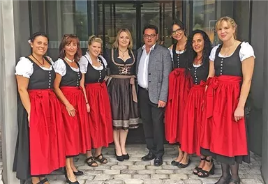 Eine Gruppe von Personen in traditioneller bayerischer Tracht steht vor einem Gebäude. Sie tragen schwarze Blusen und rote Röcke und lächeln in die Kamera.