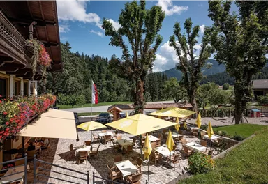 Ein einladendes Restaurant mit sonnigen Terrassen und gelben Sonnenschirmen. Im Hintergrund sind grüne Bäume und Berge zu sehen.