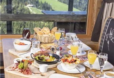 A rich breakfast buffet with various foods and drinks on a table. In the background, the green mountains and a clear sky can be seen.